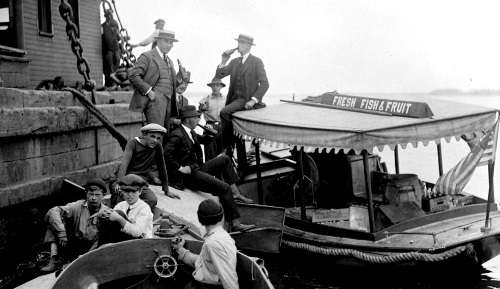 Boat with sign "Fresh Fish and Fruit" delivers bottled drinks to men on pier (possibly Prohibition selling illegal alcohol). Photo courtesy Boston Public Library, Leslie Jones Collection.