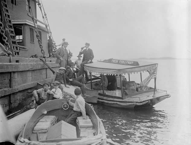 Boat with sign "Fresh Fish and Fruit" delivers bottled drinks to men on a Boston Harbor pier (probably selling illegal alcohol). Photo courtesy Boston Public Library, Leslie Jones Collection.